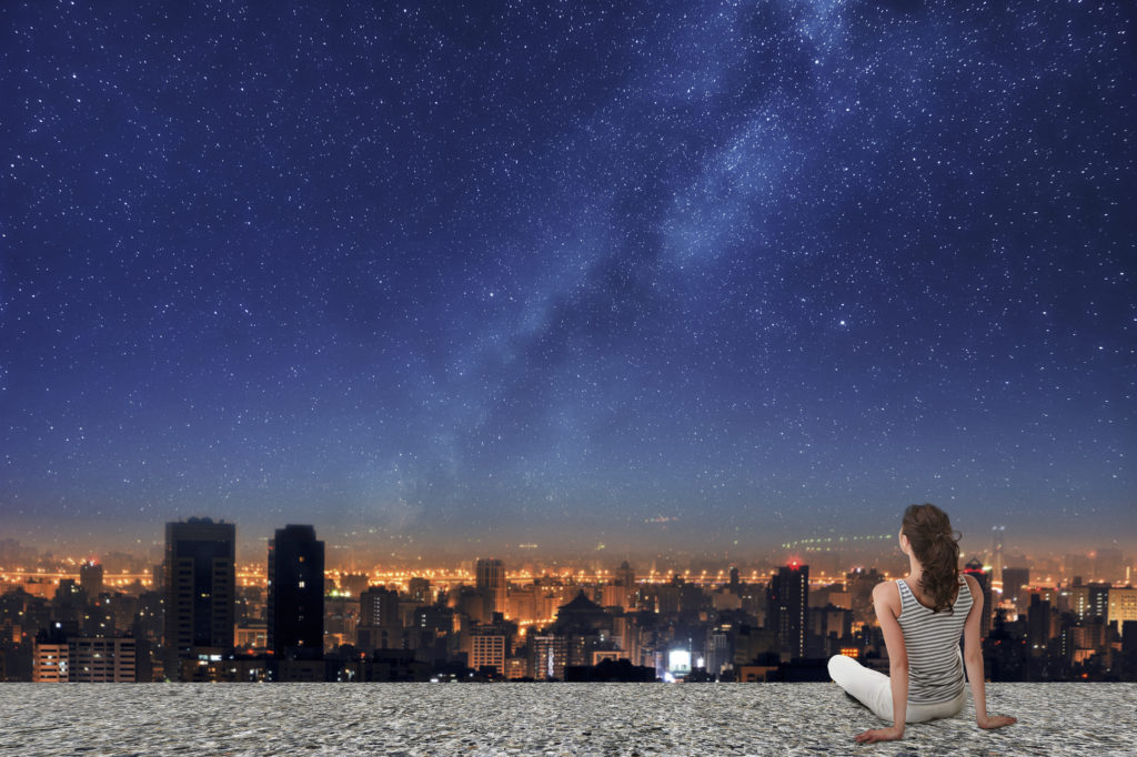 Asian woman sitting on roof, back to camera, and looking on the night city under starry sky.